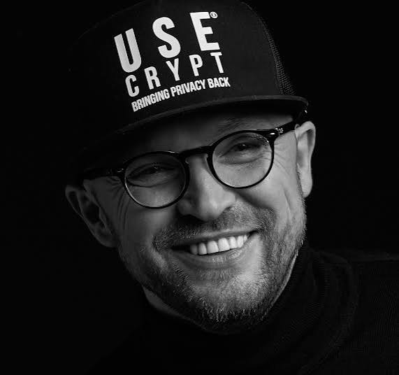 Paweł Makowski smiling, wearing glasses and a black UseCrypt cap.